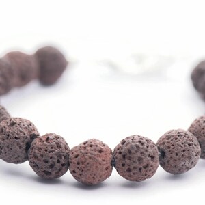 Puede incluir: Un brazalete hecho de cuentas de piedra de lava marr&oacute;n oscuro y porosa. Las cuentas redondas tienen una textura rugosa. El brazalete est&aacute; ensartado en un cord&oacute;n blanco y se muestra sobre un fondo blanco. Este brazalete de piedra natural es una joya.