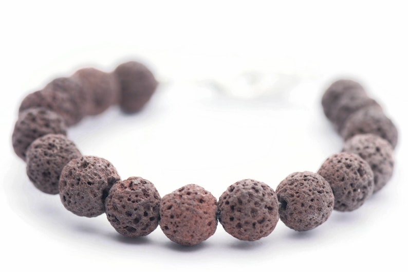 Puede incluir: Un brazalete hecho de cuentas de piedra de lava marr&oacute;n oscuro y porosa. Las cuentas redondas tienen una textura rugosa. El brazalete est&aacute; ensartado en un cord&oacute;n blanco y se muestra sobre un fondo blanco. Este brazalete de piedra natural es una joya.