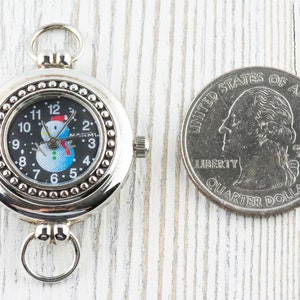 May include: A silver pocket watch with a snowman design on the face. The watch is about the size of a US quarter.