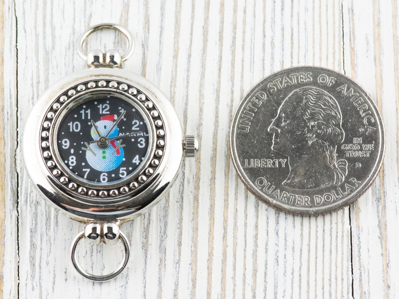 May include: A silver pocket watch with a snowman design on the face. The watch is about the size of a US quarter.