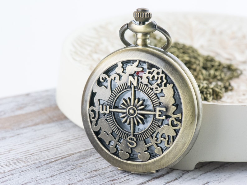 Op de afbeelding: Een zakhorloge in vintage stijl met een kompas-wijzerplaat en decoratieve uitsnijdingen rond de rand. Het horloge is gemaakt van messing en heeft een ketting.