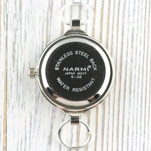 Op de afbeelding: Een zilverkleurig zakhorloge met een zwarte wijzerplaat. Het horloge heeft een ronde vorm met een roestvrijstalen achterkant en de tekst "NARMI JAPAN MOVT S-36 WATER RESISTANT". Het heeft een kleine kroon en twee ringbevestigingen.