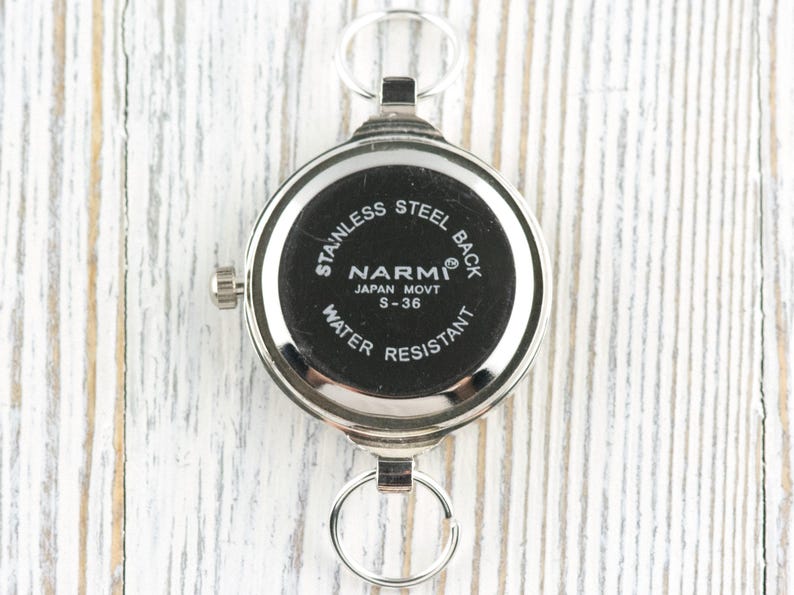 Op de afbeelding: Een zilverkleurig zakhorloge met een zwarte wijzerplaat. Het horloge heeft een ronde vorm met een roestvrijstalen achterkant en de tekst "NARMI JAPAN MOVT S-36 WATER RESISTANT". Het heeft een kleine kroon en twee ringbevestigingen.