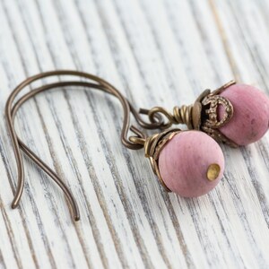 K&ouml;nnte beinhalten: Ein Paar handgefertigte Ohrringe mit rosa Holzperlen und antiken bronzefarbenen Metallakzenten. Die Ohrringe haben ein einfaches Haken-Design und sind vor einem wei&szlig;en Holzgrund platziert.