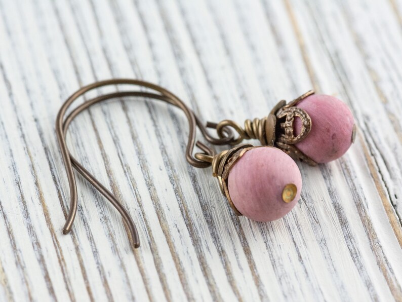 K&ouml;nnte beinhalten: Ein Paar handgefertigte Ohrringe mit rosa Holzperlen und antiken bronzefarbenen Metallakzenten. Die Ohrringe haben ein einfaches Haken-Design und sind vor einem wei&szlig;en Holzgrund platziert.