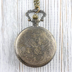 May include: Antique bronze pocket watch with a detailed floral design on the cover. The watch hangs from a matching chain. The watch case is round and features an ornate pattern of leaves and flowers.