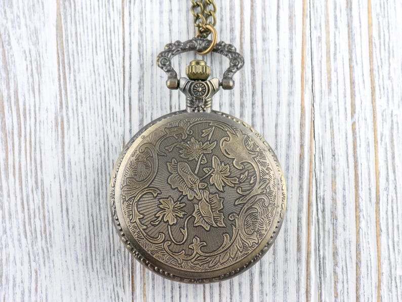 May include: Antique bronze pocket watch with a detailed floral design on the cover. The watch hangs from a matching chain. The watch case is round and features an ornate pattern of leaves and flowers.