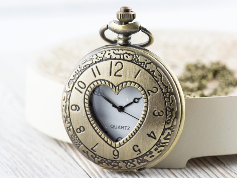 Pode incluir: Um rel&oacute;gio de bolso dourado com uma janela em forma de cora&ccedil;&atilde;o que revela o mostrador branco do rel&oacute;gio. O rel&oacute;gio tem marcas de n&uacute;meros romanos e a palavra "QUARTZ" impressa no mostrador.