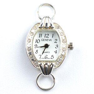 May include: Silver-toned Geneva quartz watch with a white face and black hands. The watch has a decorative oval bezel encrusted with small, clear crystals. The watch features a ring at the top and bottom for attachment.