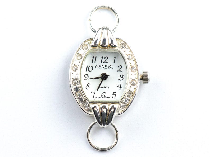May include: Silver-toned Geneva quartz watch with a white face and black hands. The watch has a decorative oval bezel encrusted with small, clear crystals. The watch features a ring at the top and bottom for attachment.