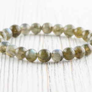 Puede incluir: Un brazalete de cuentas redondas de labradorita pulida. Las cuentas son principalmente de color verde oliva con destellos iridiscentes azules y grises. El brazalete est&aacute; ensartado en una banda el&aacute;stica.