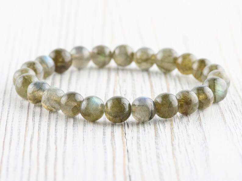 Puede incluir: Un brazalete de cuentas redondas de labradorita pulida. Las cuentas son principalmente de color verde oliva con destellos iridiscentes azules y grises. El brazalete est&aacute; ensartado en una banda el&aacute;stica.
