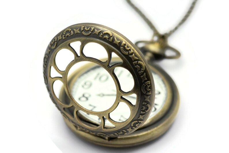 K&ouml;nnte beinhalten: Eine goldfarbene Taschenuhr mit einem verzierten, offenen Design. Die Uhr hat ein wei&szlig;es Zifferblatt mit schwarzen r&ouml;mischen Ziffern f&uuml;r die Stunden und schwarzen Zeigern.