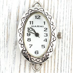 May include: Oval-shaped watch face with a silver-colored filigree border. The white face displays black numerals and hands, with the brand name "NARMI" at the top. A small crown is visible on the side.