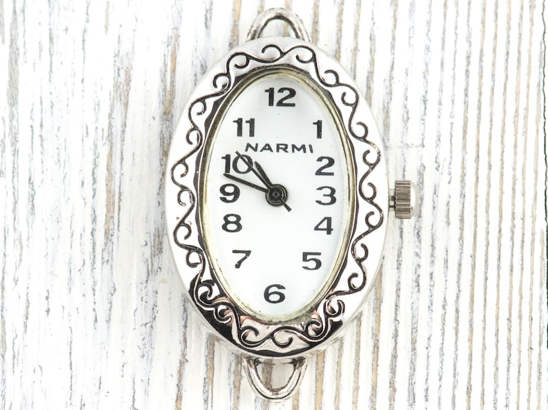 May include: An oval watch face with a silver filigree border. The white face has black numerals and hands, with the brand name "NARMI". A small crown is on the side. The watch face is a watch component.