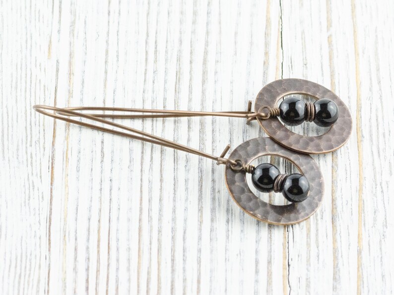 Peut inclure: Boucles d'oreilles artisanales avec des pendentifs ovales en cuivre martel&eacute;. Chaque pendentif contient deux perles noires. Les boucles d'oreilles ont un design simple et &eacute;l&eacute;gant, parfait pour un usage quotidien. Elles sont pr&eacute;sent&eacute;es sur une surface en bois blanc.