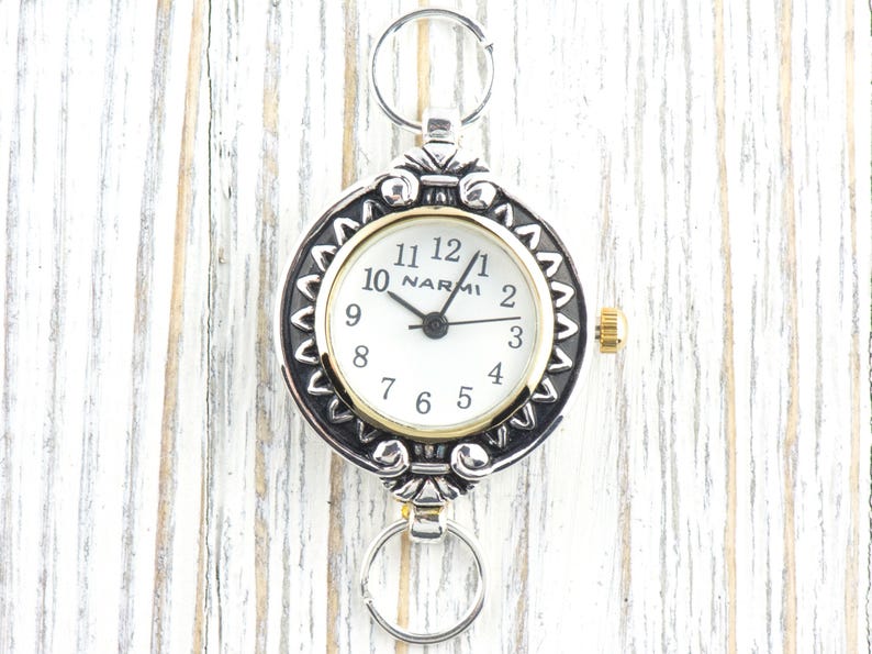 Pu&ograve; includere: Orologio da tasca color argento con quadrante bianco e dettagli dorati. L'orologio presenta il marchio "NARMI" e una lunetta decorativa. Include portachiavi nella parte superiore e inferiore per il fissaggio.