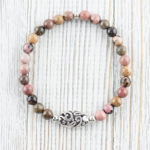 Puede incluir: Un brazalete de piedras preciosas rosa, marr&oacute;n y gris con un dije de plata. El brazalete est&aacute; hecho de cuentas redondas y tiene un cierre de plata.