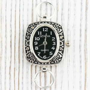 K&ouml;nnte beinhalten: Eine silberne Taschenuhr mit schwarzem Zifferblatt und r&ouml;mischen Ziffern f&uuml;r die Stunden. Die Uhr hat ein quadratisches Geh&auml;use mit einem verzierten Design und eine kleine Schlaufe oben zum Anbringen einer Kette.