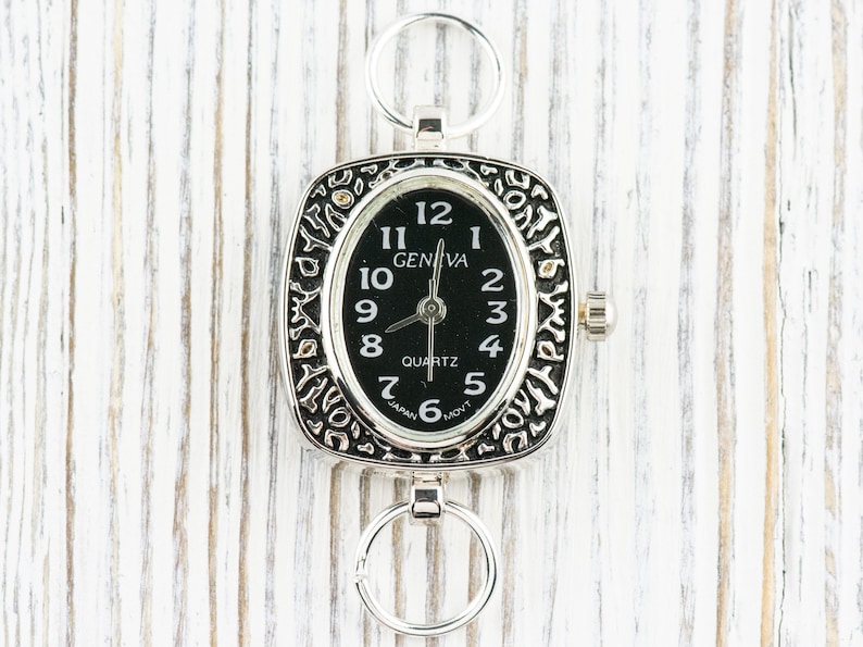 K&ouml;nnte beinhalten: Eine silberne Taschenuhr mit schwarzem Zifferblatt und r&ouml;mischen Ziffern f&uuml;r die Stunden. Die Uhr hat ein quadratisches Geh&auml;use mit einem verzierten Design und eine kleine Schlaufe oben zum Anbringen einer Kette.
