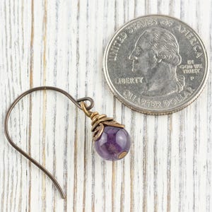 K&ouml;nnte beinhalten: Ein einzelner Ohrring mit einem dunklen Metallhaken und einer lila Edelsteinperle. Die Perle hat eine tiefviolette Farbe und ist mit einer goldfarbenen Metallkappe akzentuiert. Eine US-Vierteldollarm&uuml;nze liegt neben dem Ohrring zum Gr&ouml;&szlig;envergleich.