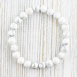 Pode incluir: Um bracelete de contas de howlite brancas com forma redonda. O bracelete est&aacute; colocado sobre uma superf&iacute;cie de madeira branca.