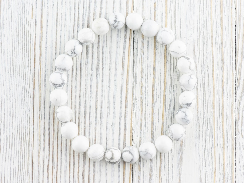 Pode incluir: Um bracelete de contas de howlite brancas com forma redonda. O bracelete est&aacute; colocado sobre uma superf&iacute;cie de madeira branca.