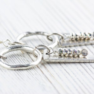 May include: A pair of silver hoop earrings with a silver dangle charm. The dangle charm features a row of silver beads and a silver disc.