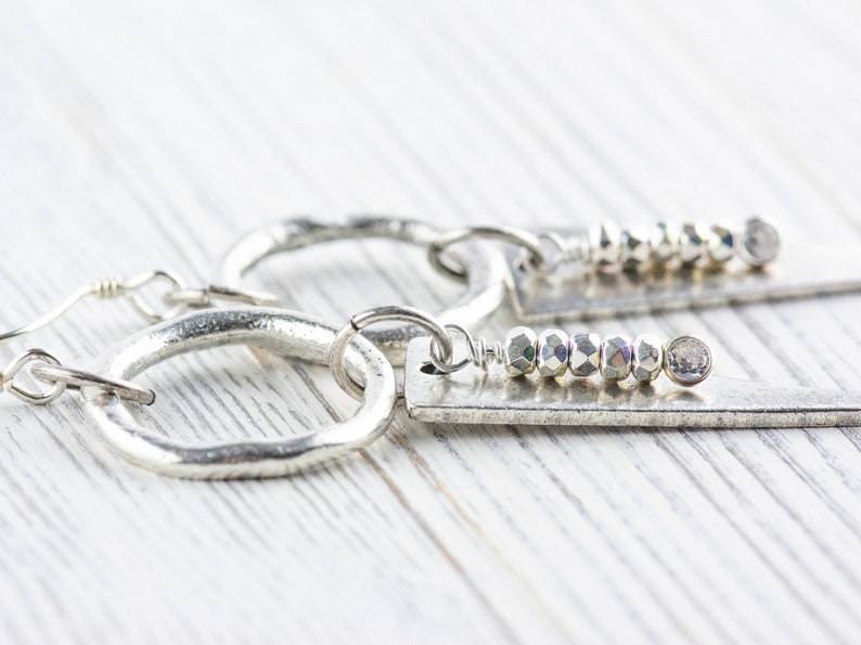 May include: A pair of silver hoop earrings with a silver dangle charm. The dangle charm features a row of silver beads and a silver disc.