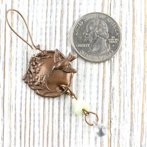 May include: A bronze-colored charm with a bird and grape design, with a quarter for size comparison. The charm has a loop at the top and a dangling chain with a light green bead and a clear teardrop-shaped bead.