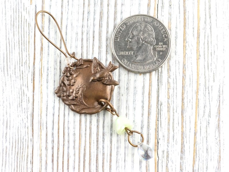 May include: A bronze-colored charm with a bird and grape design, with a quarter for size comparison. The charm has a loop at the top and a dangling chain with a light green bead and a clear teardrop-shaped bead.