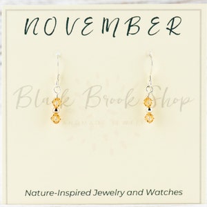 May include: A pair of silver dangle earrings with two amber-coloured crystal beads on each earring. The earrings are displayed on a white card with the text "NOVEMBER" at the top and "Black Brook Shop Handmade Jewelry" in the middle. The bottom of the card reads "Nature-Inspired Jewellery and Watches".
