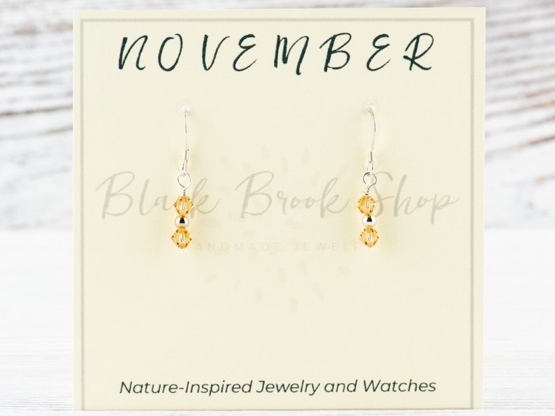 May include: A pair of silver dangle earrings with two amber-coloured crystal beads on each earring. The earrings are displayed on a white card with the text "NOVEMBER" at the top and "Black Brook Shop Handmade Jewelry" in the middle. The bottom of the card reads "Nature-Inspired Jewellery and Watches".