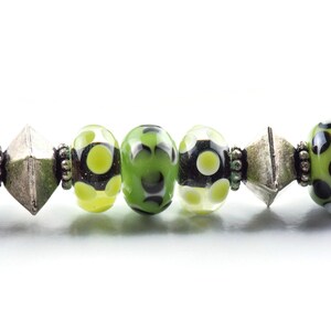 May include: A close-up of a green and black patterned glass bead bracelet with silver spacers. The bracelet features a variety of green and black beads with white polka dots.