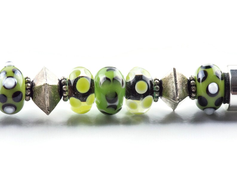 May include: A close-up of a green and black patterned glass bead bracelet with silver spacers. The bracelet features a variety of green and black beads with white polka dots.