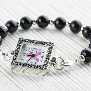 Op de afbeelding: Een vierkante Geneva quartz horloge met een roze lint ontwerp op de wijzerplaat, bevestigd aan een armband van zwarte kralen. Het horloge heeft een zilverkleurig frame en sluiting.