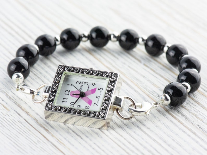 Op de afbeelding: Een vierkante Geneva quartz horloge met een roze lint ontwerp op de wijzerplaat, bevestigd aan een armband van zwarte kralen. Het horloge heeft een zilverkleurig frame en sluiting.