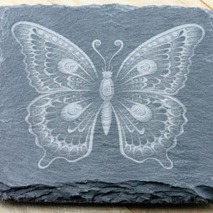Puede incluir: Un posavasos de pizarra gris con un dise&ntilde;o de mariposa blanca grabada. La mariposa tiene detalles intrincados y una apariencia delicada.