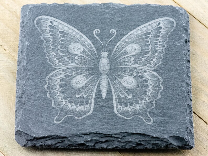Puede incluir: Un posavasos de pizarra gris con un dise&ntilde;o de mariposa blanca grabada. La mariposa tiene detalles intrincados y una apariencia delicada.