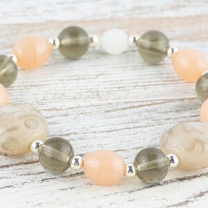 May include: A bracelet made with light peach, grey, and white beads. The beads are strung on a silver chain.