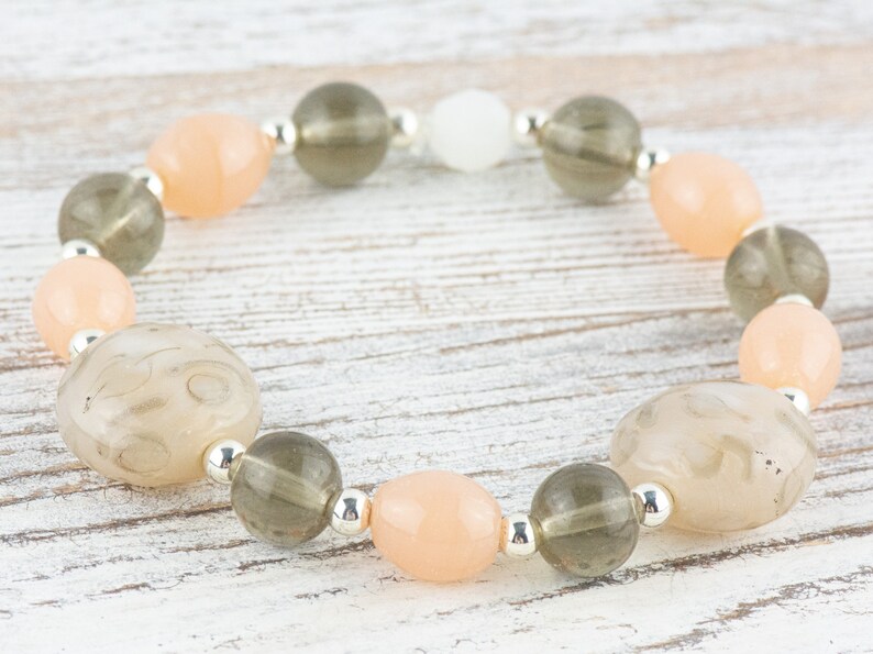 May include: A bracelet made with light peach, grey, and white beads. The beads are strung on a silver chain.