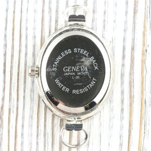 May include: A silver-toned watch back with a black face. The watch back has the text "STAINLESS STEEL BACK", "GENEVA JAPAN MOVT L-20", and "WATER RESISTANT". The watch has a loop at the bottom and a crown on the side.