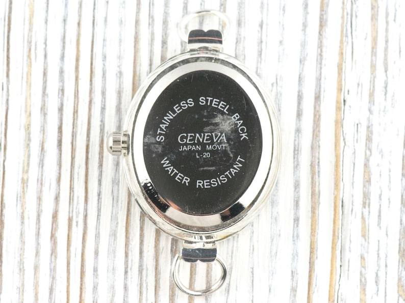 May include: A silver-toned watch back with a black face. The watch back has the text "STAINLESS STEEL BACK", "GENEVA JAPAN MOVT L-20", and "WATER RESISTANT". The watch has a loop at the bottom and a crown on the side.