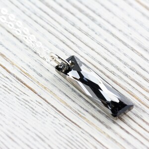 Pode incluir: Um colar de corrente de prata com um pingente de cristal preto. O pingente tem uma forma retangular alongada com facetas que captam a luz.