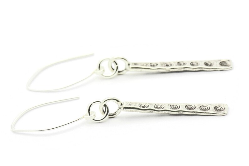 May include: A pair of silver dangle earrings with a textured bar design. The bar has a repeating pattern of small circles.