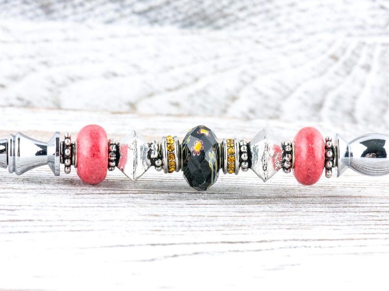 May include: A silver-toned bead bracelet with a variety of decorative beads. The bracelet features red, black, and clear beads, along with silver and gold-colored accents. The beads are arranged in a symmetrical pattern.