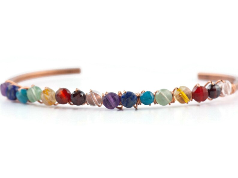 K&ouml;nnte beinhalten: Ein mit Kupferdraht umwickeltes Armband mit einem Regenbogen aus Edelsteinen, darunter Amethyst, Lapislazuli, T&uuml;rkis und klarer Quarz.