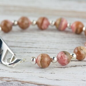 May include: A silver bracelet with a personalized tag that reads "ASTHMA" and pink and brown gemstone beads.