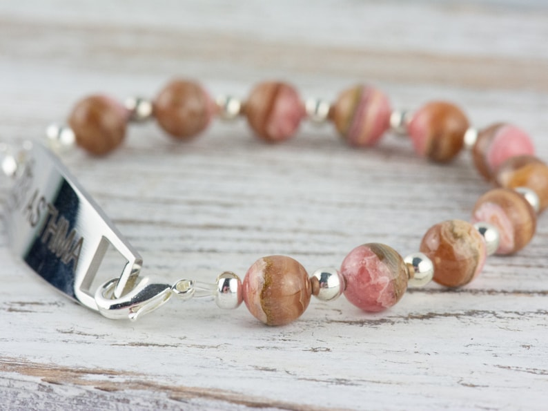 May include: A silver bracelet with a personalized tag that reads "ASTHMA" and pink and brown gemstone beads.