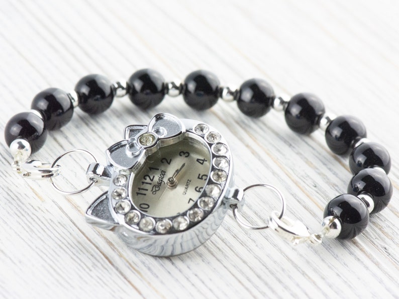 May include: A silver watch with a black cat face design and rhinestones around the face. The watch is attached to a black beaded bracelet with a silver clasp.